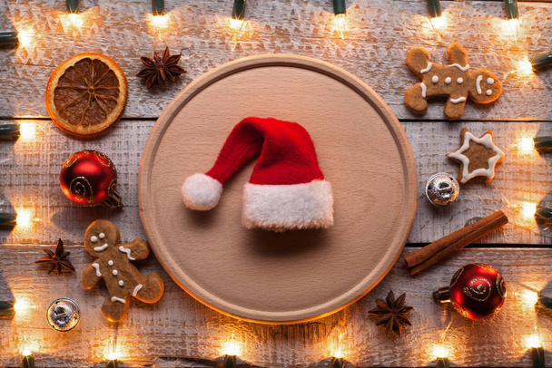 Préparation pour Noël et la période des fêtes - avec des décorations de Noël, biscuits et ingrédients sur la table - chapeau de Père Noël au centre sur une assiette en bois
 - Photo, image