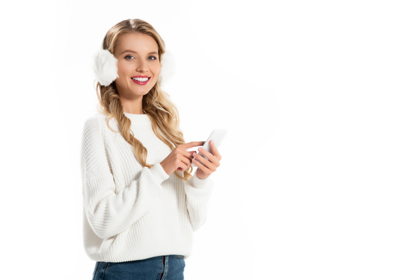 attractive smiling girl in earmuffs using smartphone isolated on white - Fotografie, Obrázek
