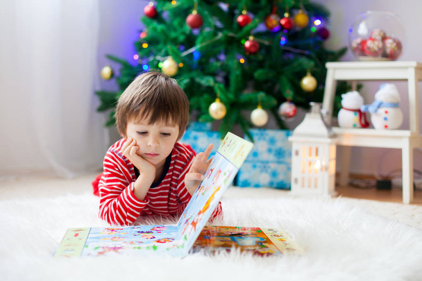 Cute boy, reading a book in front of Christmas tree at home - Fotografie, Obrázek