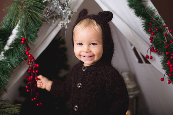 Cute toddler baby boy in handmade knitted bear costume, playing in teepee, decorated for Christmas, studio shot on brown background - Foto, immagini