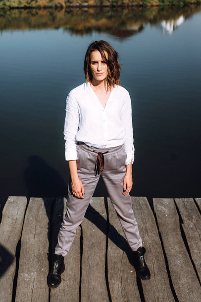 白いシャツとグレーのズボンの桟橋は、川と紅葉葉の背景の上に立ってポーズでスタイリッシュな女性. - 写真・画像