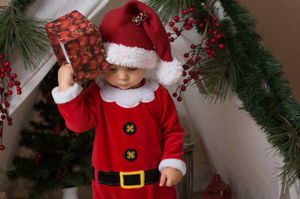 Чарівний маленький малюк хлопчик, одягнений в костюм Клауса, грає вдома перед тепі, прикрашений на Різдво
 - Фото, зображення