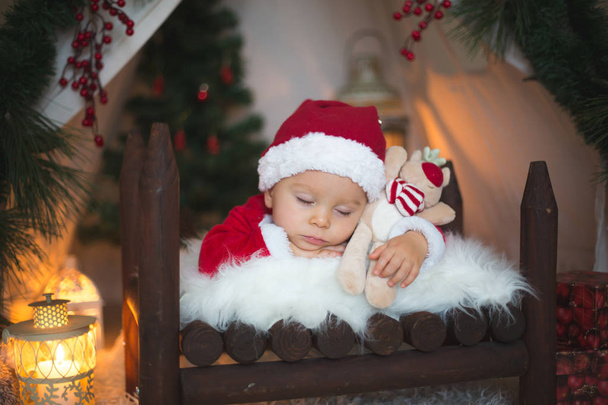 Adorable niño pequeño bebé vestido con traje de canta claus, durmiendo en la cama de bebé en frente de tipi decorado para Navidad
 - Foto, imagen