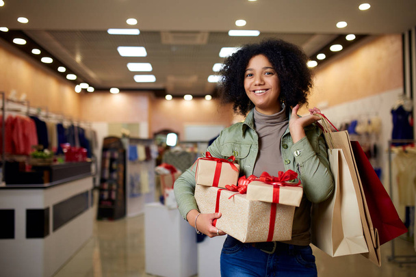 African american girl shopping gifts in mall on christmas sale. New year holidays concept. Smiling attractive mixed racial woman with colorful paper gift boxes wearing christmas hat in store or shop. - Fotografie, Obrázek