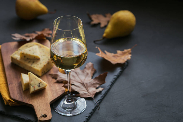 A glass of white wine was served with cheese in a cutting board on dark background. Autumn picnic with cheese, wine and dry leaves in rustic style. - 写真・画像