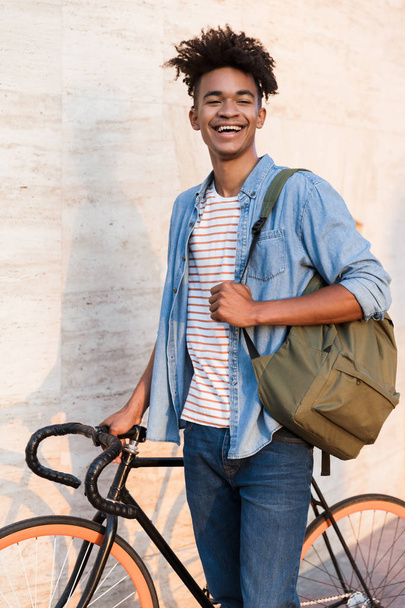Image of happy laughing young african guy walking with bicycle outdoors on the street. - 写真・画像