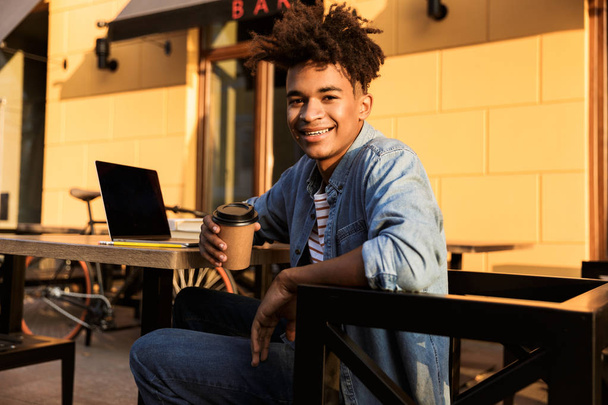 Happy young african man sitting at the cafe outdoors, holding coffee cup - Foto, Imagen