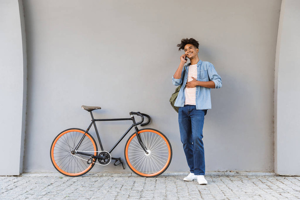 Image of happy young african guy walking outdoors with bicycle talking by mobile phone. - Valokuva, kuva