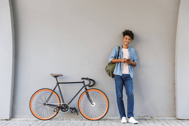 Image of happy young african guy walking outdoors with bicycle chatting by mobile phone. - Foto, imagen