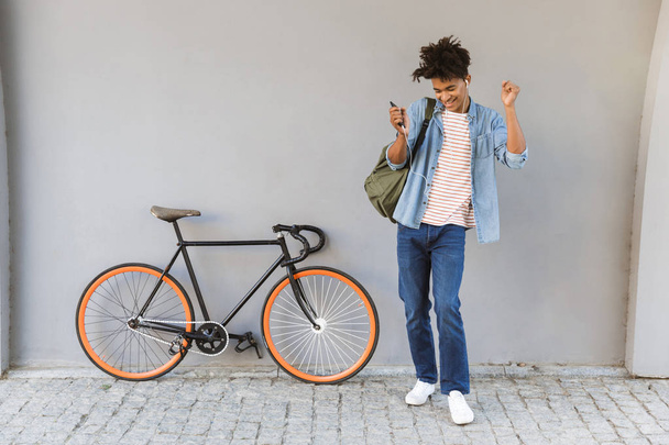 Image of happy young african guy walking outdoors with bicycle listening music chatting by mobile phone. - Foto, immagini