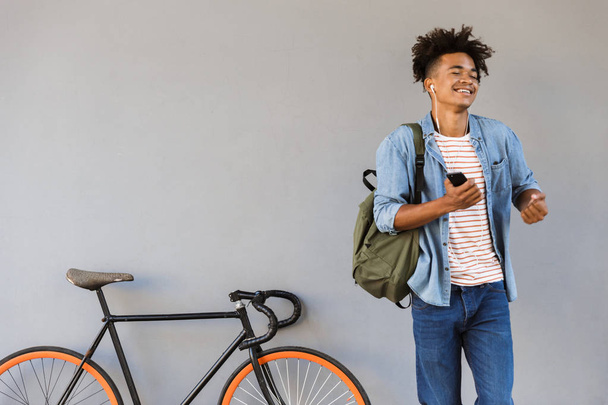 Image of cheerful excited young african guy walking outdoors with bicycle listening music holding mobile phone. - Foto, Bild