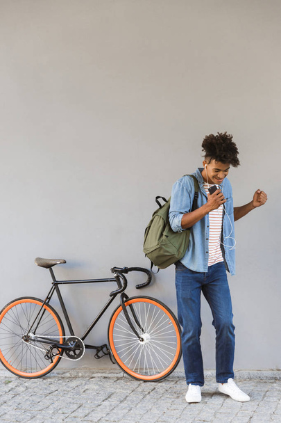 Image of cheerful excited young african guy walking outdoors with bicycle listening music holding mobile phone. - Fotó, kép