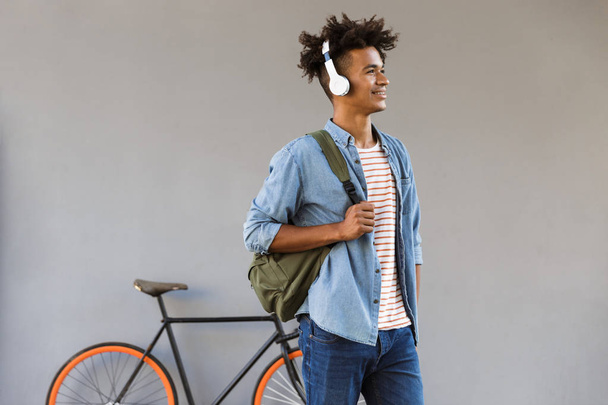 Image of cheerful young african guy walking outdoors with bicycle listening music holding backpack. - Foto, imagen