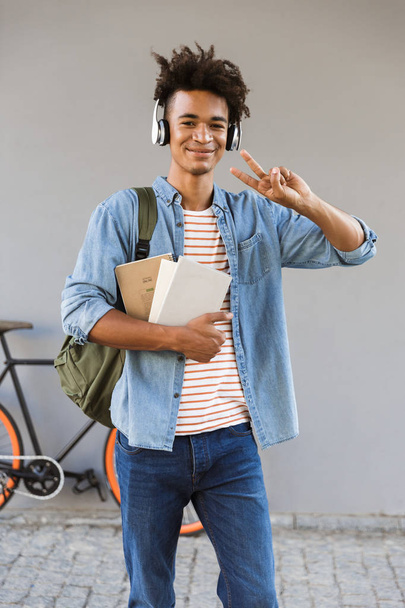 Smiling young african man with backpack outdoors, standing with bicycle, listening to music with headphones, holding books - Photo, image