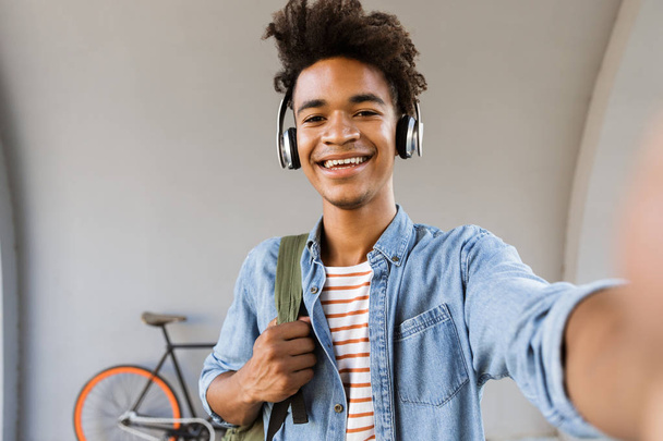 Smiling young african man with backpack outdoors, standing with bicycle, listening to music with headphones, taking a selfie - Foto, Bild