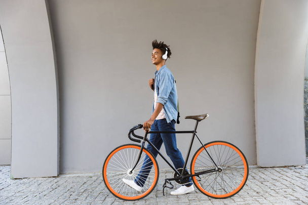Smiling young african man outdoors, walking with bicycle, listening to music with headphones - Zdjęcie, obraz
