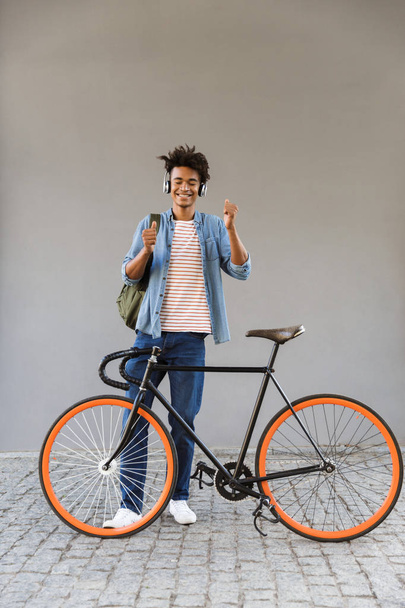Cheerful young african man outdoors, standing with bicycle, listening to music with headphones - Fotoğraf, Görsel