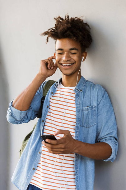 Image of cheerful excited young african guy walking outdoors listening music holding mobile phone. - Photo, Image