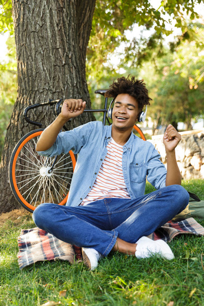 Excited young african teenager with bicycle outdoors, sitting on grass, listening to music with headphones, having fun - Fotografie, Obrázek