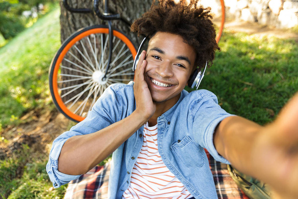 Happy young african teenager with bicycle outdoors, sitting on grass, listening to music with headphones, taking a selfie - Фото, зображення