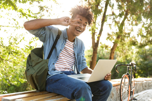 Image of handsome emotional young african guy in park outdoors using laptop computer showing peace gesture. - Foto, Imagen