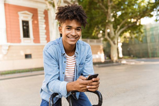 Smiling young african man outdoors, riding on a bicycle, holding mobile phone - Фото, зображення