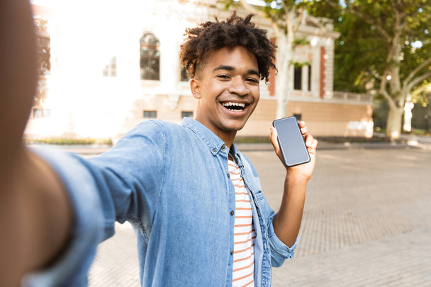 アフリカ青年屋外で、selfie を取って、空白の画面携帯電話を示す笑みを浮かべてください。 - 写真・画像