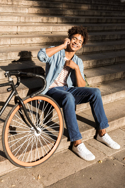 Image of cheerful young african guy sitting outdoors with bicycle on the street on steps talking by phone. - Photo, Image