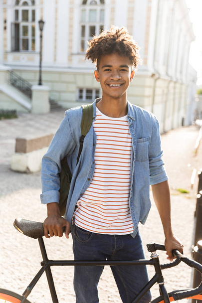 Image of cheerful excited young african guy walking outdoors with bicycle on the street. - 写真・画像
