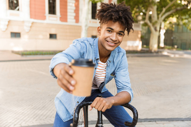 Image of cheerful excited young african guy walking outdoors with bicycle holding coffee. - Photo, Image