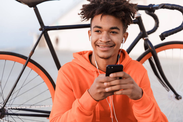 Image of a young sports guy outdoors on the beach with bicycle using mobile phone listening music. - Photo, Image