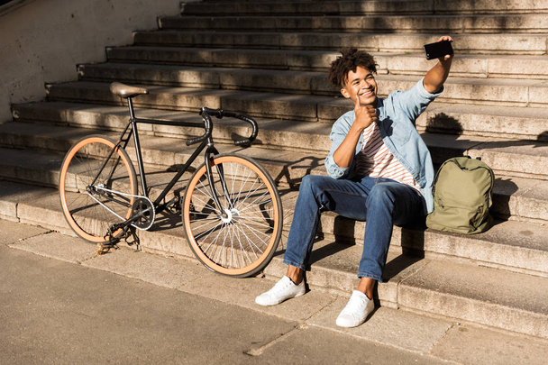 Smiling young african man with backpack outdoors, sitting on stairs with bicycle, taking a selfie with mobile phone - Zdjęcie, obraz