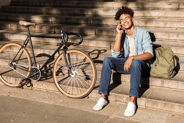 Smiling young african man with backpack outdoors, sitting on stairs with bicycle, talking on mobile phone - Foto, Imagem