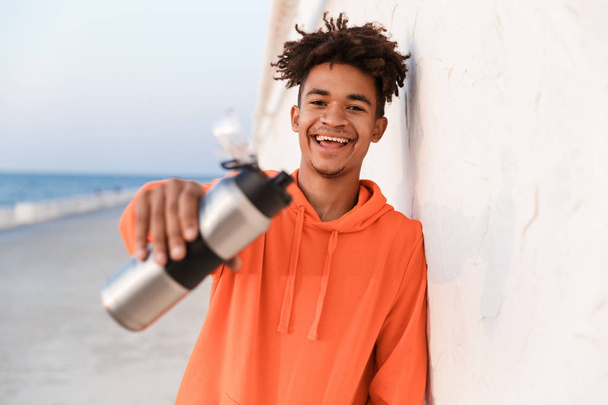 Image of a young sports guy outdoors on the beach drinking water holding bottle. - Foto, imagen