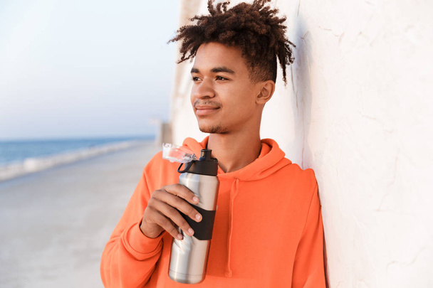 Image of a young sports guy outdoors on the beach drinking water holding bottle. - Photo, image