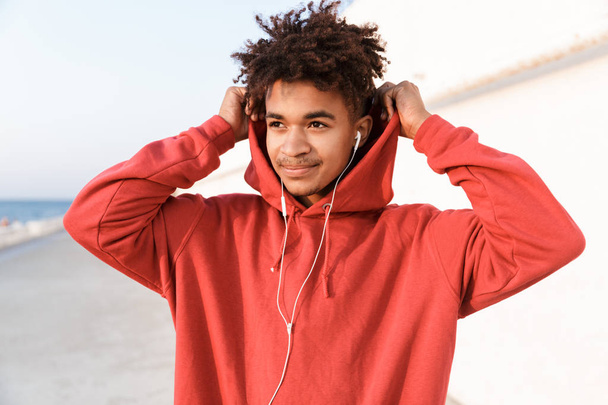 Close up of a handsome young african teenager dressed in hoodie standing at the beach, listening to music with earphones - 写真・画像