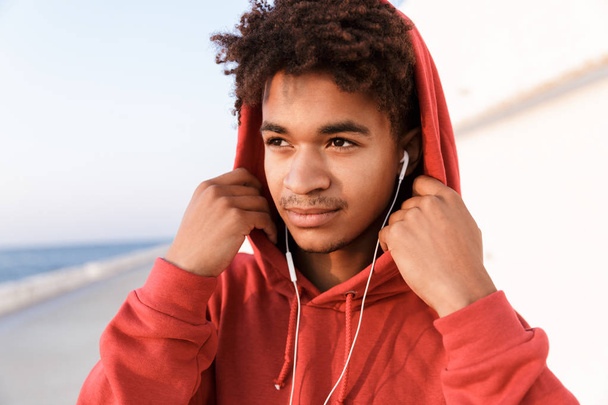 Close up of a young african teenager dressed in hoodie standing at the beach, listening to music with earphones - Foto, Imagen