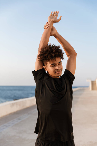 Image of handsome young guy sportsman make stretching exercises outdoors on the beach. - Foto, afbeelding