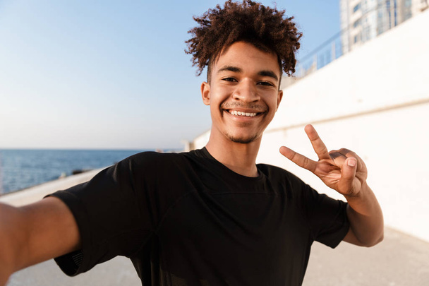 Sonriente joven africano adolescente deportista de pie en la playa, tomando una selfie con teléfono móvil, mostrando gesto de paz
 - Foto, imagen