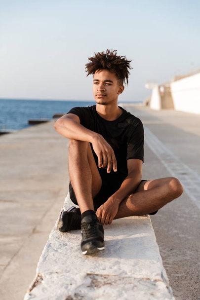 Image of concentrated young guy sportsman sitting outdoors on the beach. - Φωτογραφία, εικόνα