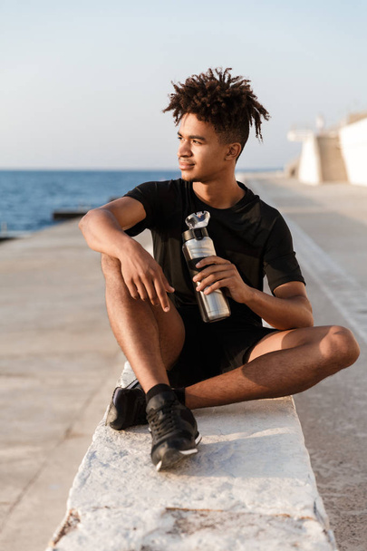 Image of handsome young guy sportsman sitting outdoors on the beach drinking water. - Photo, Image