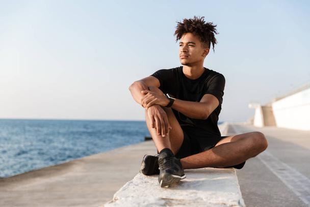Image of concentrated young guy sportsman sitting outdoors on the beach. - Φωτογραφία, εικόνα