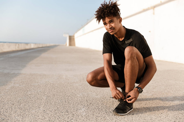 Strong young african man dressed in sportswear tying his shoelace at the beach - Foto, imagen
