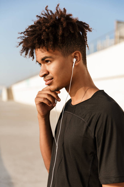 Image of a young guy sportsman walking outdoors on the beach listening music with earphones. - Photo, Image