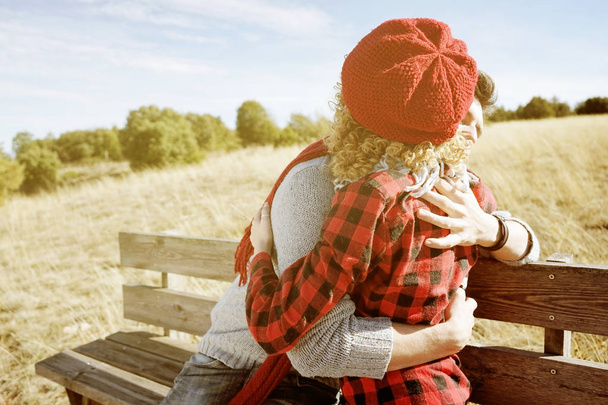 gelukkige jonge paar verliefd knuffelen en zonnebaden op houten bankje op weide op zonnige herfstdag - Foto, afbeelding