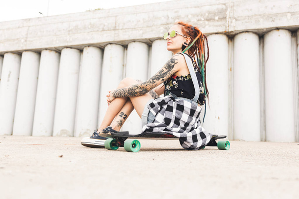 young girl with tattoo and dreadlocks sitting on a longboard on the background of concrete structure - Foto, Imagen
