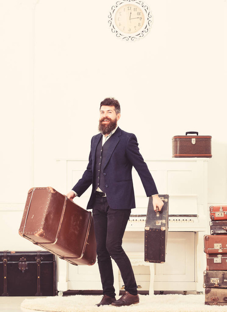 Macho attractive, elegant on smiling face carries vintage suitcases. Man with beard and mustache wearing classic suit delivers luggage, luxury white interior background. Butler and service concept - Photo, Image