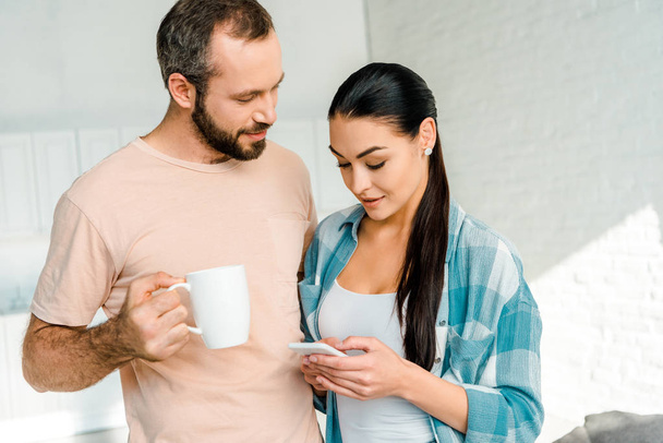 mooie brunette vrouw gebruikend smartphone terwijl echtgenoot holding kopje koffie thuis - Foto, afbeelding