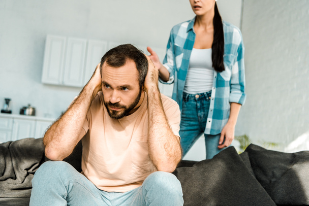 estresado marido en primer plano cubriendo las orejas con las manos y tener una discusión con la esposa en casa
 - Foto, Imagen