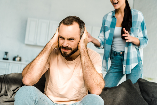 frustrated husband on foreground covering ears with hands and having argument with wife at home - Photo, Image